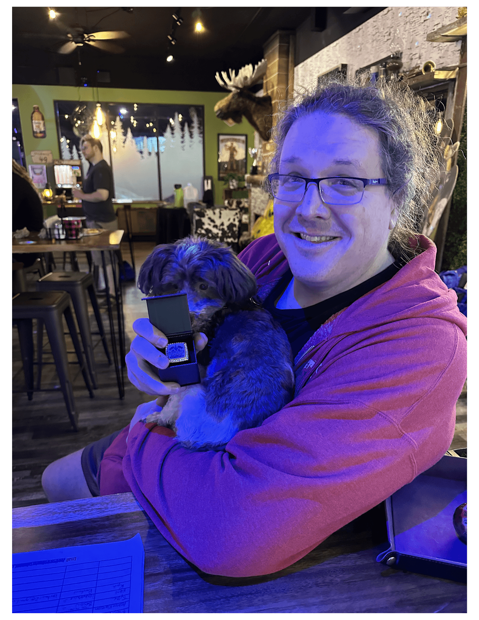 A smiling man in a red hoodie holds a small dog and a ring box.