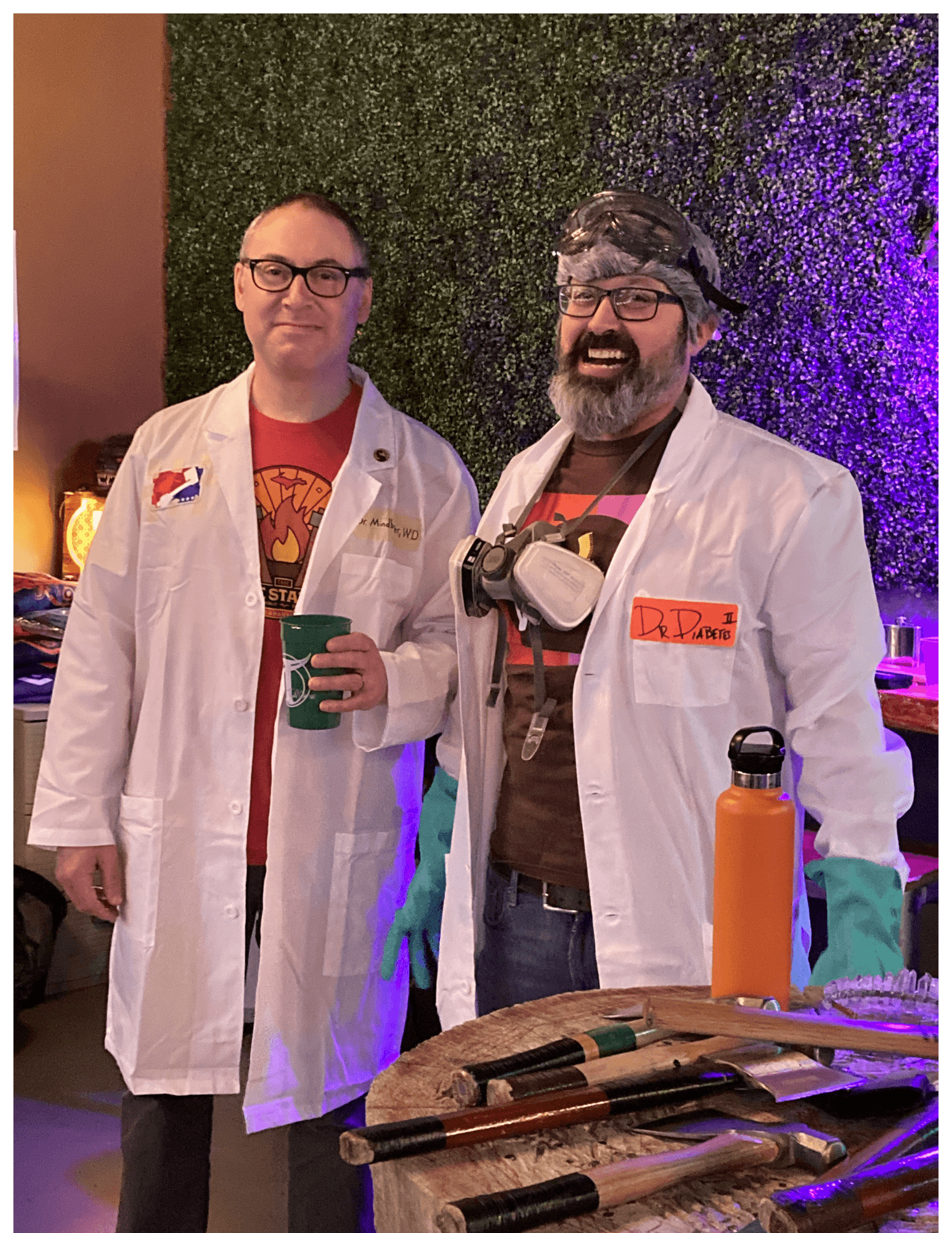 Two men in lab coats and safety gear stand behind a table of hand tools.