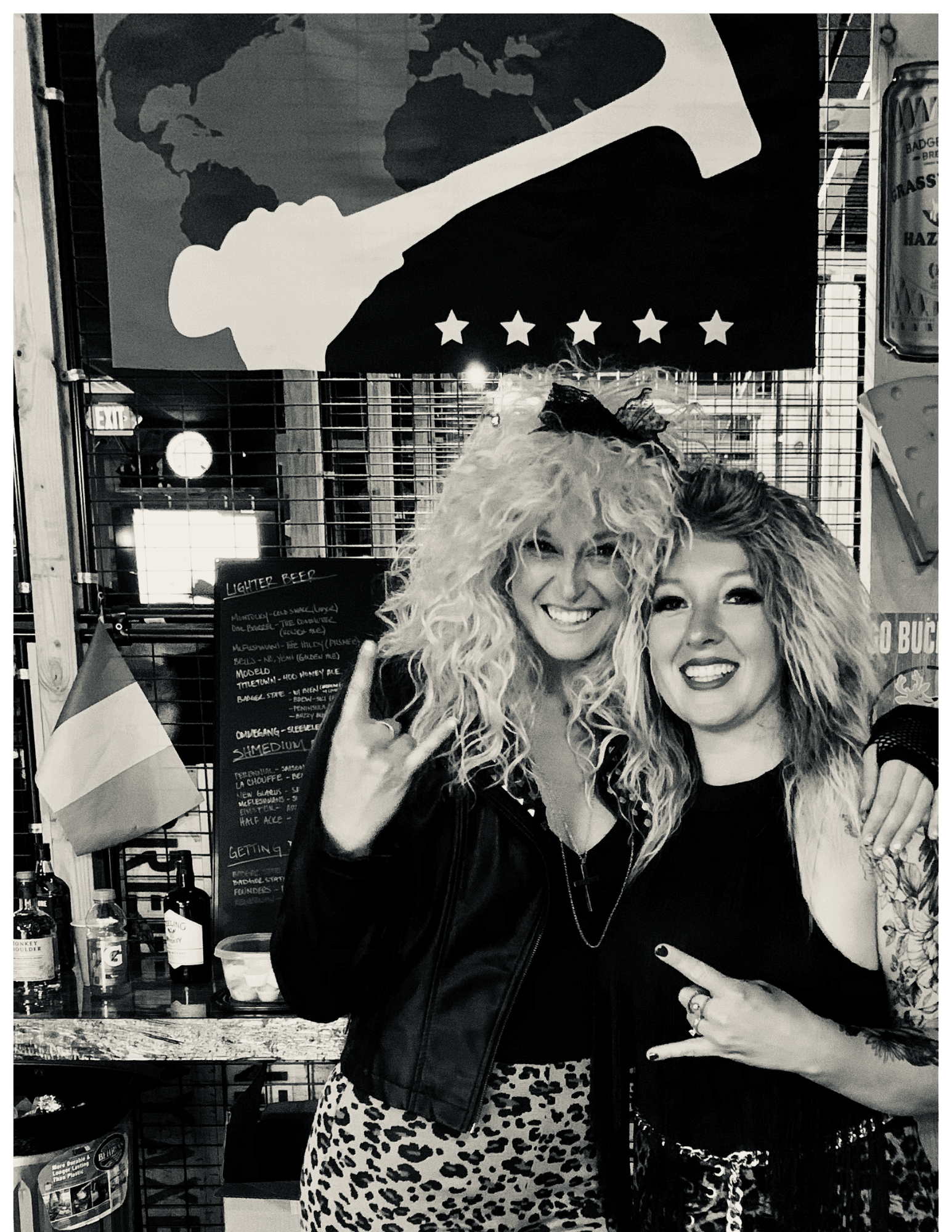 Two smiling women with big 80s hair making rock hand signs in a bar.