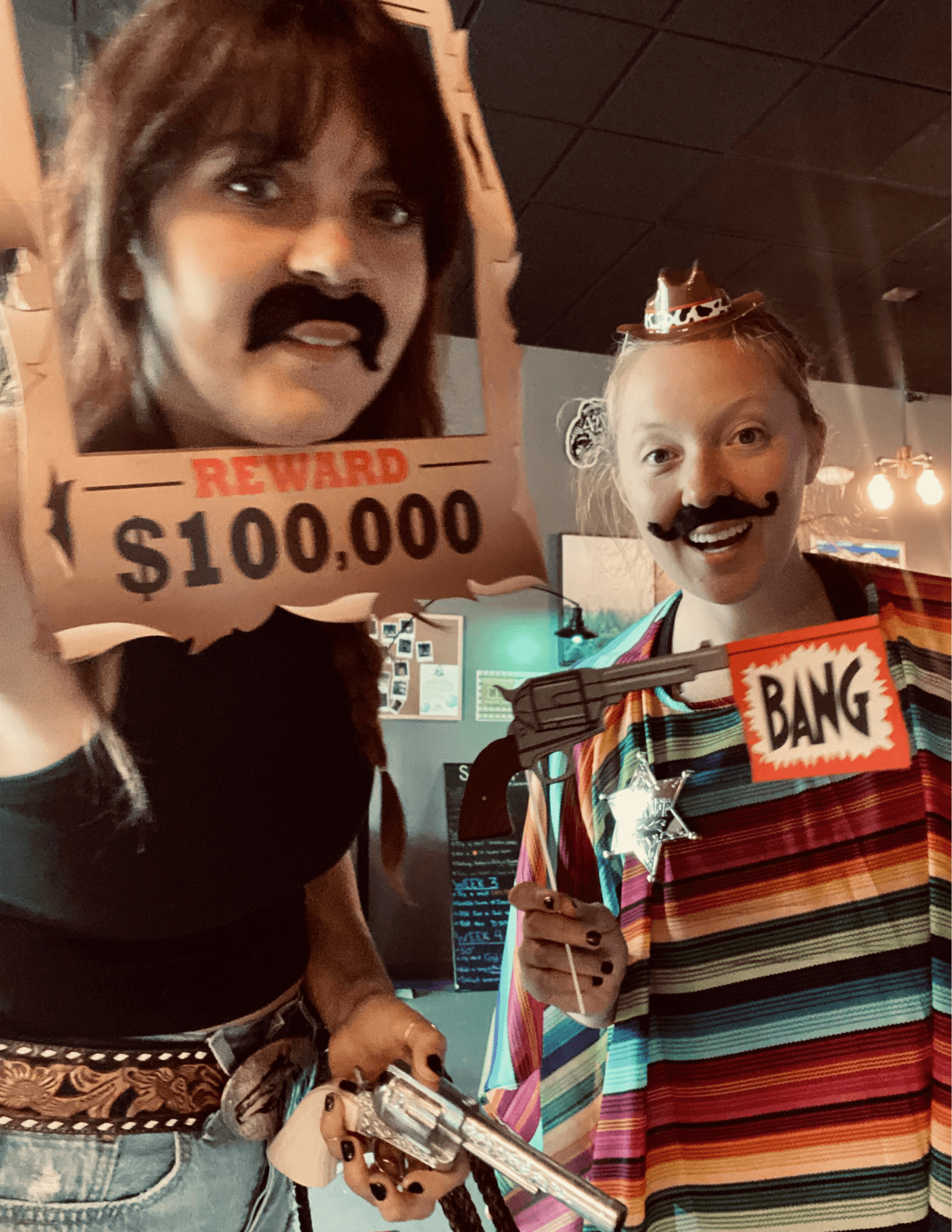 Two women wearing fake mustaches pose with a wanted poster frame and a toy gun.