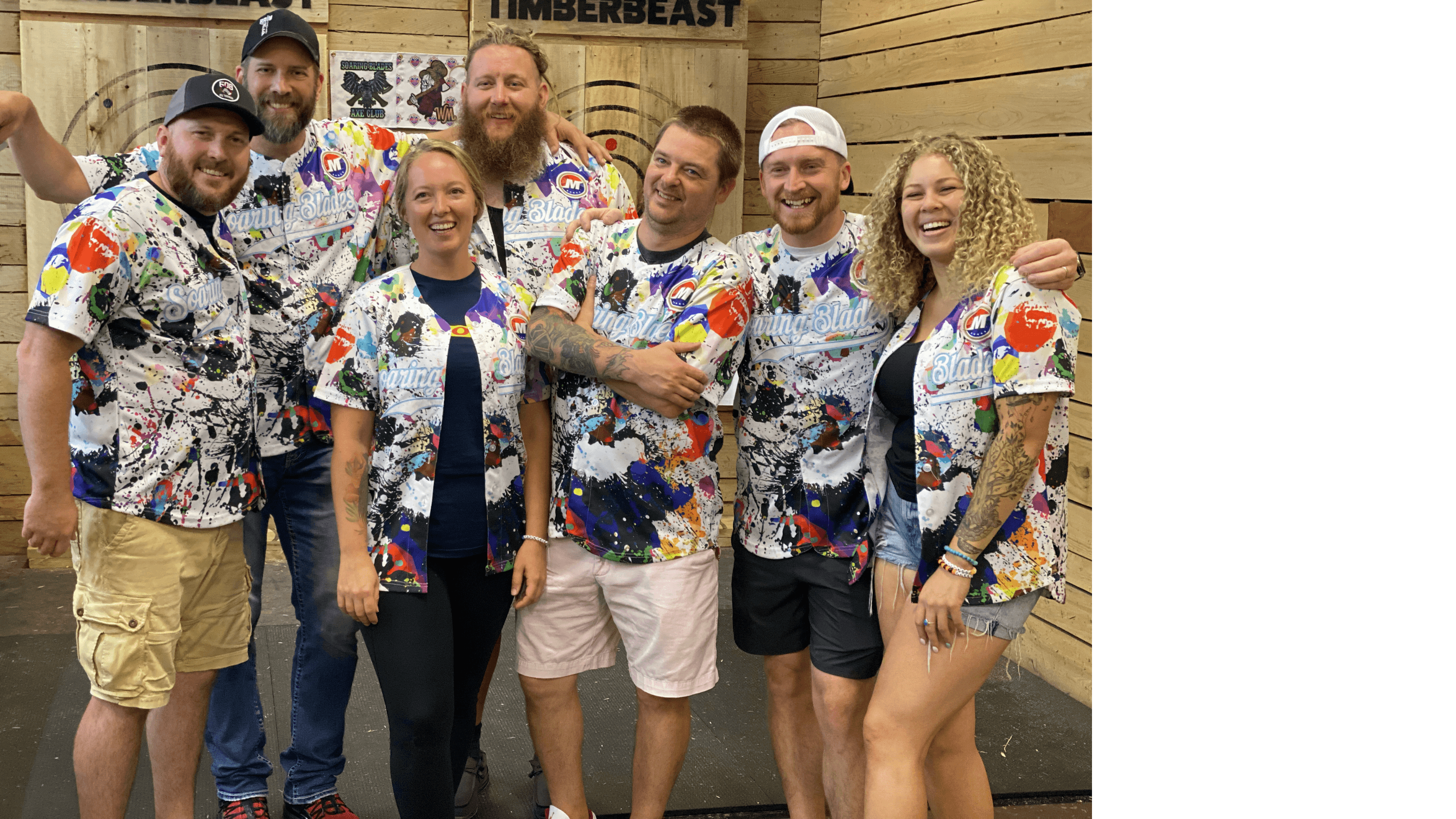 Six people in paint-splattered Soaring Blades jerseys smile at an axe-throwing venue.