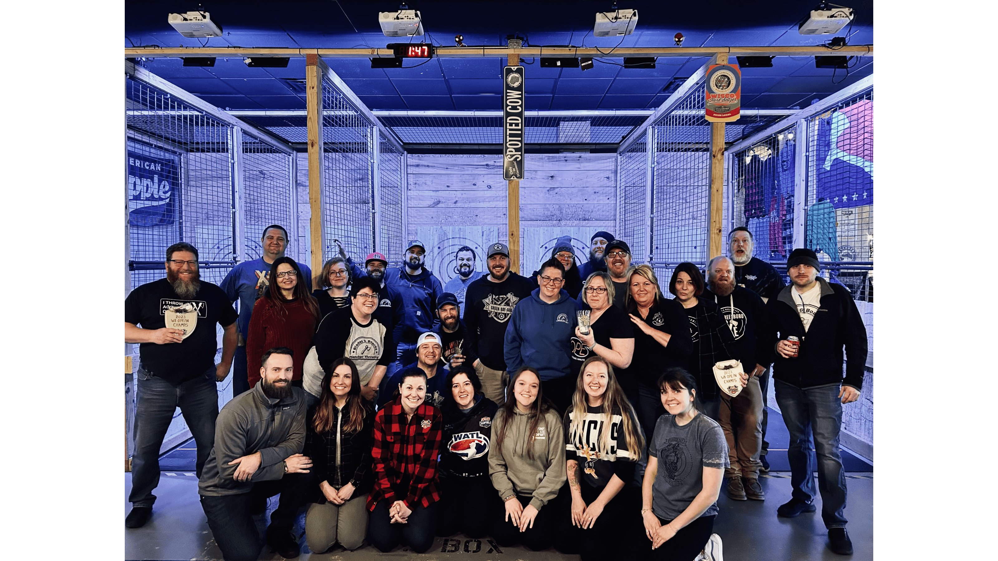 Large group of people posing with trophies inside an indoor axe-throwing facility with wooden targets.