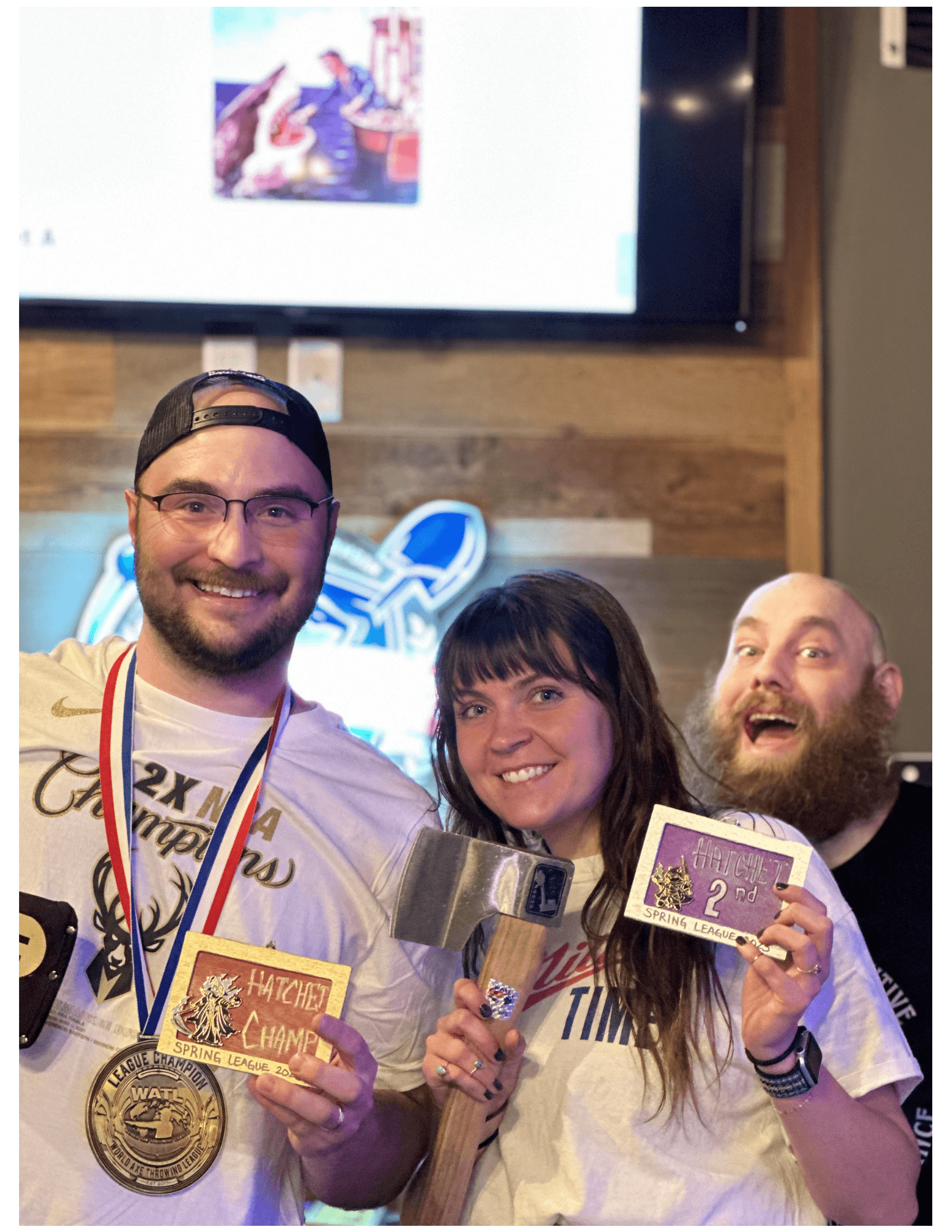Smiling man and woman hold hatchet throwing awards and a hatchet while being photobombed.