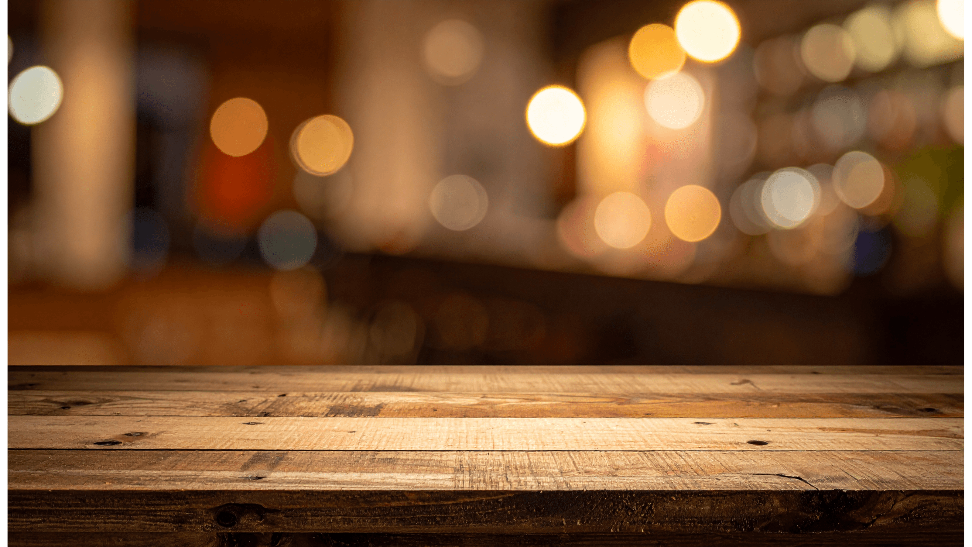 Rustic wooden tabletop in front of a blurred bar interior with warm, glowing bokeh lights.